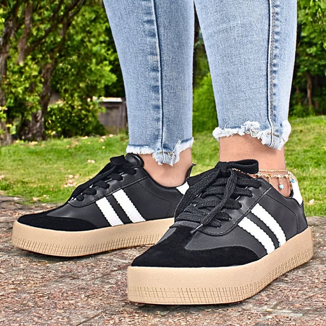 Modelo femenino caminando con Zapatilla Suelaviva Vento en negro desde la rodilla hacia abajo, diseño retro atemporal con cuero sintético y ante sintético, suela de goma spansor beige claro, bandas blancas laterales, ideal para un estilo deportivo y sofisticado.