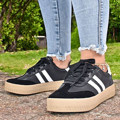 Modelo femenino caminando con Zapatilla Suelaviva Vento en negro desde la rodilla hacia abajo, diseño retro atemporal con cuero sintético y ante sintético, suela de goma spansor beige claro, bandas blancas laterales, ideal para un estilo deportivo y sofisticado.