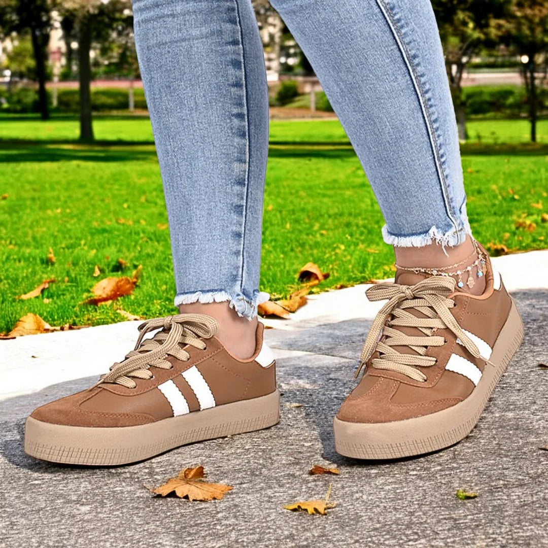 Modelo femenino en movimiento con Zapatilla Suelaviva Vento en marrón desde la rodilla hacia abajo, diseño retro y versátil con ante sintético y cuero sintético, suela de goma beige claro para adherencia, bandas blancas laterales, ideal para un look activo y moderno.