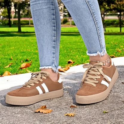 Modelo femenino en movimiento con Zapatilla Suelaviva Vento en marrón desde la rodilla hacia abajo, diseño retro y versátil con ante sintético y cuero sintético, suela de goma beige claro para adherencia, bandas blancas laterales, ideal para un look activo y moderno.